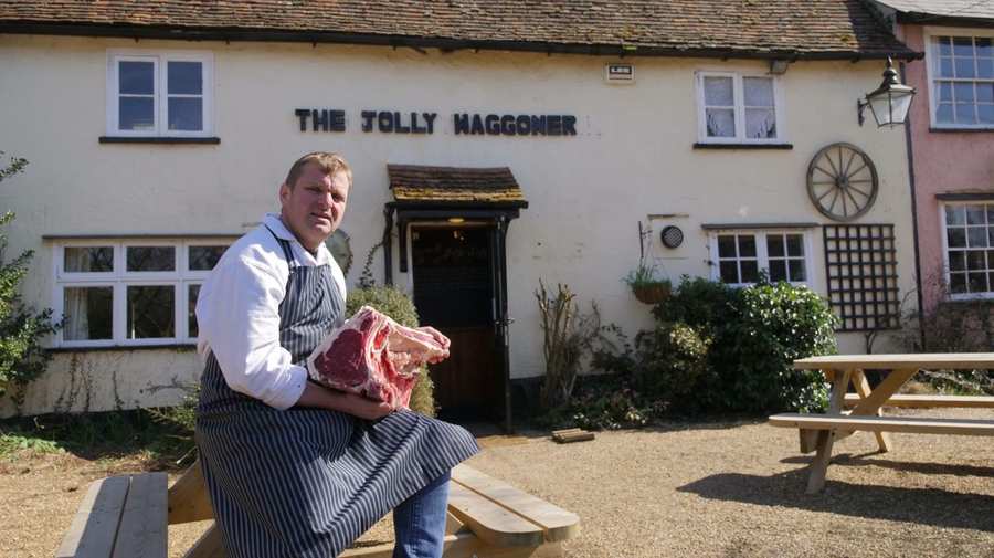 A butcher in a striped apron sitting outside The Jolly Waggoner pub, holding a côte de boeuf